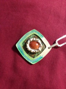 A snapped a goldstone cabachon into the bezel. I used two part epoxy to attach the bezel to the copper, and then the copper to the frame. Solder would not work, because one would lose the patina. Once two-part epoxy is thoroughly dry, it will stand up to normal wear. Attach a pinch bail. The piece is complete!
